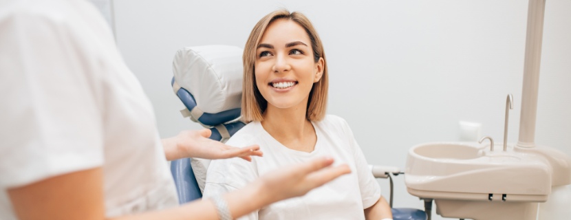 woman with blonde cropped hair sat in dental chair, smiling with a straight, white smile and looking up at a dentist who is explaining a procedure