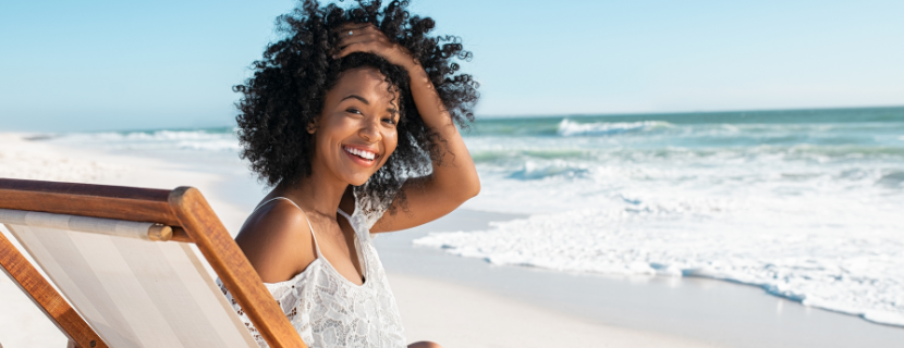 a black woman sitting on the beach by the sea, looking over at the camera with her hand in her hair and smiling with a bright white smile at the camera