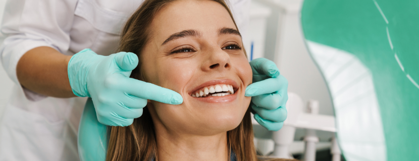 pretty girl smiling in a dentist's chair with her new dazzling smile whilst a dentist points at her teeth with pale green gloves on.