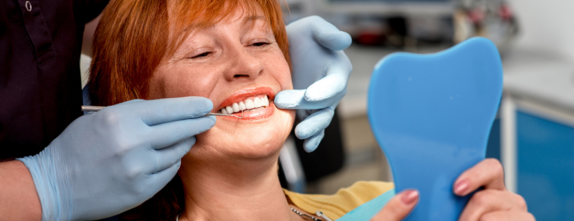 ginger older woman smiling as she holds up a cosmetic mirror, looking at her white smile in a dentist chair as a dentist's hands wearing blue gloves points at her teeth 