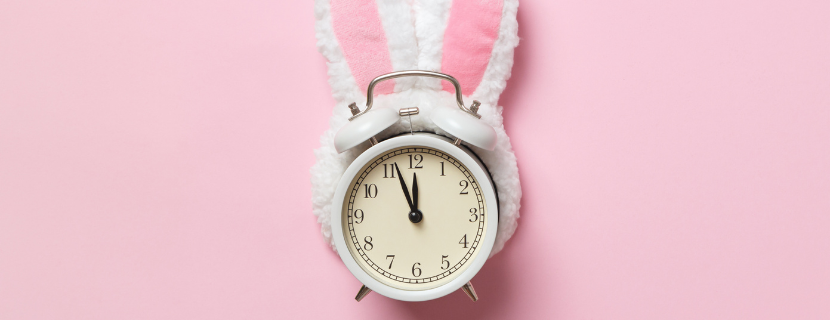 image of an alarm clock against a pink background with white fluffy bunny ears wrapped around it to symbolise Easter